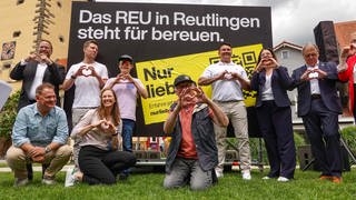 Die Organisatoren der Werbekampagne des Stadtmarketings posieren vor einem Werbeplakat und formen dabei ein Herz mit den Händen. Auf dem Plakat ist zu lesen: "Das REU in Reutlingen steht für bereuen".