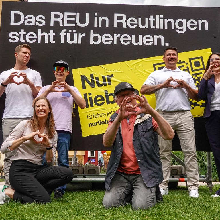 Die Organisatoren der Werbekampagne des Stadtmarketings posieren vor einem Werbeplakat und formen dabei ein Herz mit den Händen. Auf dem Plakat ist zu lesen: "Das REU in Reutlingen steht für bereuen".