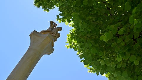 Er schleudert Blitze, der römische Gott, weit oben auf der Säule, die höher zu sein scheint als der Baum ini der rechten Bildhälfte