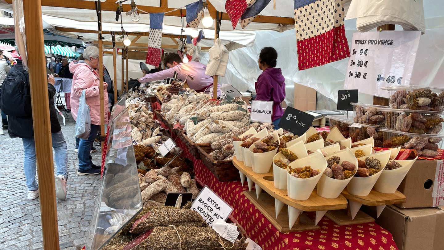 Ein Stand aus der Provence verströmt intensiven Salami-Geruch. Ein Verkäufer bedient eine Kundin. Überall hängen Frankreich-Fahnen. Beim umbrisch-provenzalischen Markt Tübingen entsteht so mit weiteren Ständen und anderen Spezialitäten ein intensiver Duft.