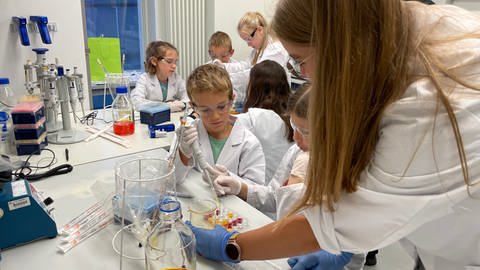 Kinder in Laborkitteln und mit Schutzhauben lernen in einem Labor der Uniklinik Tübingen pipettieren.