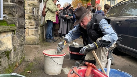 Der Künstler Gunter Demnig verlegt neue Stolpersteine in den Boden einer Tübinger Straße. Im Hintergrund stehen Menschen und schauen zu.