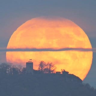 Im Vordergrund der Gipfel der Reutlinger Achalm mit der Burgruine, dahinter steigt langsam ein riesiger, tief orangener Mod auf. 