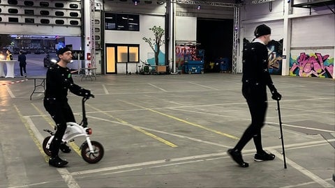 Zwei Menschen mit Laufrad und Gehstock in schwarzem Ganzkörperanzug samt kleiner Bewegungssensoren - so soll die KI menschliche Bewegungen besser verstehen.