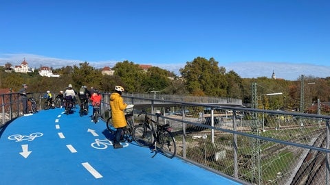 Erwachsene und Kinder genießen die Aussicht von der neuen Radbrücke West in Tübingen. Ein Gruppe Radfahrer schlängelt sich zwischen ihnen hindurch. Mit Menschen an den Seiten wird der Radweg sehr eng.