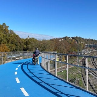 Ein älterer Herr und ein Junge stehen mit ihren Rädern auf der neuen Radbrücke West in Tübingen. Sie blicken in Richtung Schloss und Schlossberg. Zur anderen Seite liegt der Hauptbahnhof, den man ebenfalls gut beobachten kann.