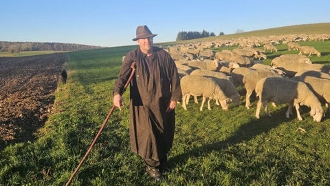 Stefan Fauser steht in Schäfertracht auf einem Feld mit seinen Schafen. Auf der Schwäbischen Alb sucht der Wanderschäfer Flächen für seine Schafe in der Nacht.