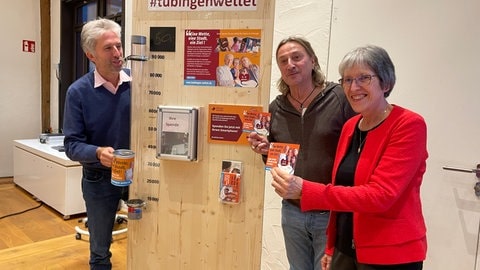 Boris Palmer hält eine Spendenbüche, Dieter Thomas Kuhn und Gisela Schneider Postkarten mit Link zum Onlinespenden hoch. Im Hintergrund die noch leere Plexiglasröhre des Spendenbarometers.