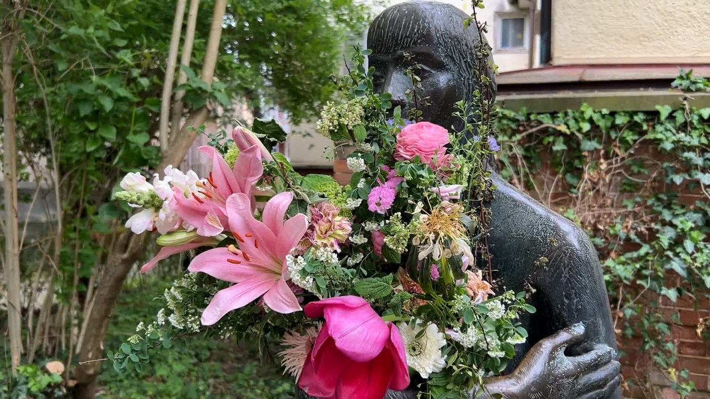 Man sieht eine Skulptur in einer Gasse in Tübingen. Sie hat einen großen Blumenstrauß in den Armen. Man nennt sie die 