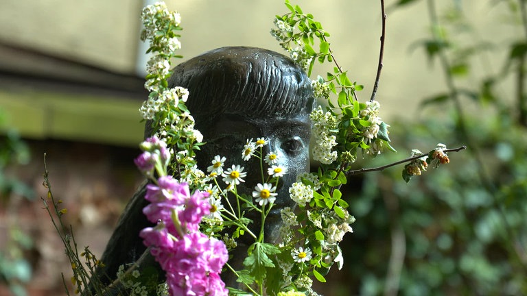 Man sieht den Kopf einer Skulptur. Man erkennt ihn nur fast, denn im Arm der Skulptur wurden Blumen abgelegt. Die Blumen sind üppig, ein ganzer Strauß verdeckt das halbe Gesicht. Die "Sitzende", so heißt die Skupltur, hat fast immer Blumen im Arm. Manchmal nur eine Handvoll, meistens aber sogar ganze Sträuße. 