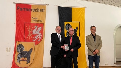 Vor einer Partnerschafts-Fahne Tomaszowski-Freudenstadt und einer Flagge des Kreis Freudenstadt (mit dem Auerhahn) stehen Landrat Rückert, Autor Herden und Lektor Kritzinger.