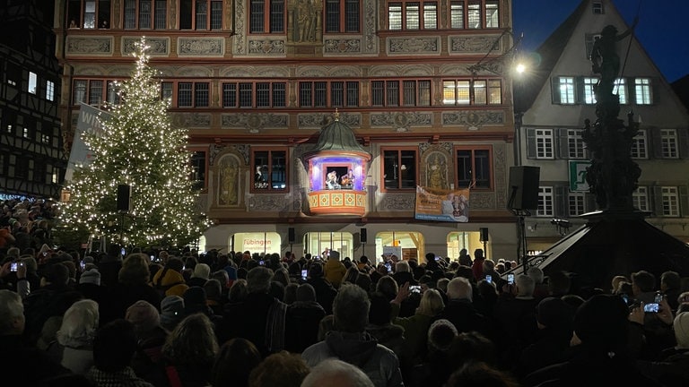 Das Tübinger Rathaus ist beim Auftritt von Dieter Thomas Kuhn beleuchtet. Davor steht ein geschmückter Weihnachtsbaum. Die Menschen auf dem Marktplatz hören zu, singen mit und applaudieren.