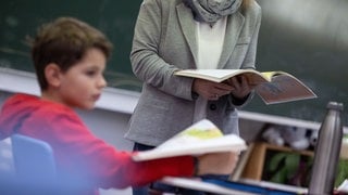 Schüler mit Lehrerin mit Buch in der Hand im Klassenzimmer: An Tübinger Schulen soll unter anderem an Schulsozialarbeit gespart werden wegen Schulden. Dagegen regt sich Protest.