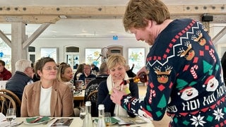 Diakon Dominik Weiß trägt einen Weihnachtspulli und schenkt bei einer Verkostung in Baiersbronn Rum in ein Glas ein. Zwei Frauen schauen ihm dabei zu. 