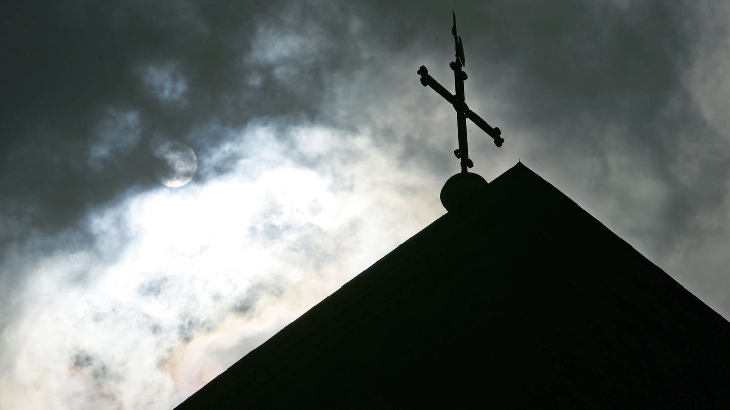 Im Gegenlicht und vor wolkenverhangenem Himmel ist eine Kirchturmspitze mit Kreuz zu sehen.