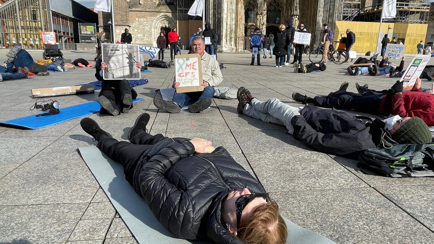 Menschen liegen auf dem Münsterplatz in Ulm, im Hintergrund das Münster. Sie demonstrieren im Liegen, um auf die Krankheit ME/CFS aufmerksam zu machen. Betroffene sind oft zu erschöpft, um das Haus zu verlassen.