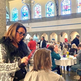 Eine Frau schneidet einer anderen in einer Kirche die Haare. Seit 20 Jahren schneidet Friseurmeisterin Maria Hartmann Gästen der Ulmer Vesperkirche kostenlos die Haare. 