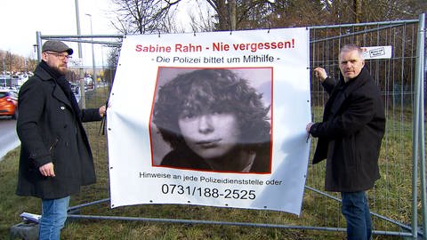 Zwei Männer befestigen ein Banner an einem Bauzaun. Der ungeklärte Mordfall Sabine Rahn beschäftigt Angehörige und Ermittler auch nach 40 Jahren noch.