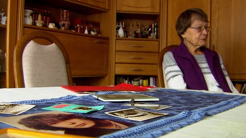 Im Vordergrund Bilder auf einem Tisch, dahinter eine ältere Frau. Der ungeklärte Mordfall Sabine Rahn beschäftigt Angehörige und Ermittler auch nach 40 Jahren noch.