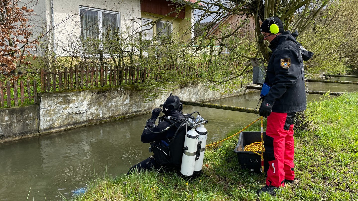 Ein Taucher sitzt am Ufer eines Bachs und macht sich zum Tauchgang bereit. Nach dem Gewaltverbrechen an einem Ehepaar in Altenstadt (Kreis Neu-Ulm) ermittelt die Kriminalpolizei weiter in alle Richtungen. Am Montag suchten Ermittler rund um das Wohnhaus der Opfer nach Spuren.