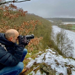 Ein Mann blickt mit einer Kamera in der Hand übers Lonetal. Tierfotograf Jochen Bayer auf dem Hubertusfelsen, wo Wanderfalken brüten. 