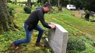 Ein Mann drückt mit Kraft gegen einen hellgrauen Grabstein auf einem Friedhof in Aalen, im Hintergrund Bäume, Gräber und Wiese.