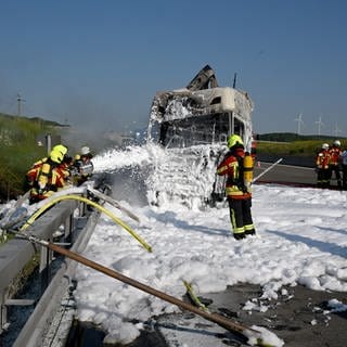 Das ausgebrannte Führungshaus eines Lkw. Feuerwehrleute stehen in einem Teich aus Löschschaum. Mehrere sprühen weiter Schaum auf den gelöschten Lkw. Im Hintegrund stehen weitere Einsatzkräfte auf der leeren A8 ind er sommerlichen Landschaft unter blauem Himmel. Sie ist zwischen Hohenstadt und Merklingen gesperrt. 