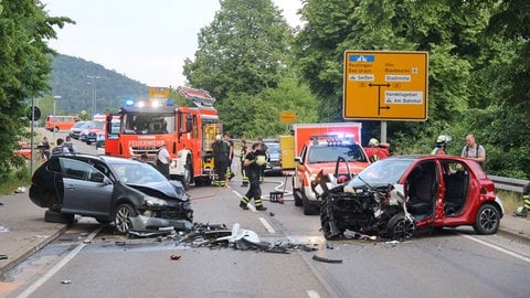 Eine Straße mit zwei an der Front völlig zerstörten Autos. Die Türen fehlen, Airbags wurden ausgelöst, es liegen Autoteile auf der Straße. Dahinter stehen Feuerwehr- und Polizeiautos sowie Einsatzkräfte. Ein Senior ist bei einem Unfall auf der B492 bei Blaubeuren in den Gegenverkehr geraten und gegen das Auto einer dreiköpfigen Familie geprallt. Unter den vier schwer Verletzten ist auch ein vierjähriges Kind.