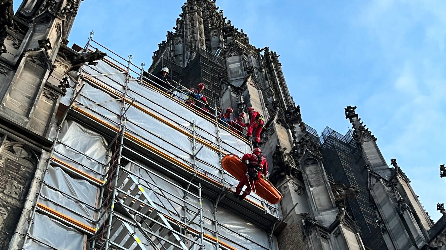 Höhenretter der Feuerwehr seilen an einem Gerüst am Ulmer Münster eine orangefarbene Trage ab. Am Münster in Ulm hat es eine Übung für Höhenrettung gegeben. An einer Trage seilten die Höhenretter eine Trage ab.