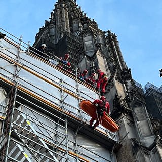 Höhenretter der Feuerwehr seilen an einem Gerüst am Ulmer Münster eine orangefarbene Trage ab. Am Münster in Ulm hat es eine Übung für Höhenrettung gegeben. An einer Trage seilten die Höhenretter eine Trage ab.