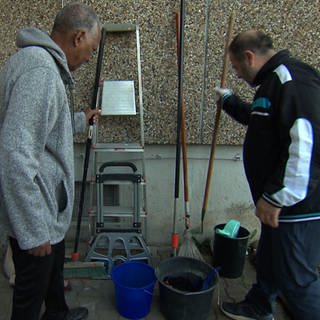 Menschen greifen nach an einer Hauswand stehenden Besen und Rechen. Rebellion mit Besenstil: Die Mieter und Mieterinnen in der Johannes-Palm-Straße 87 in Ulm greifen beherzt zu den "Waffen". Sie haben den Kampf um ihre Kehrwoche gewonnen, putzen jetzt weiter selbst und sparen sich dabei das Geld für einen externen Reinigungsservice. 