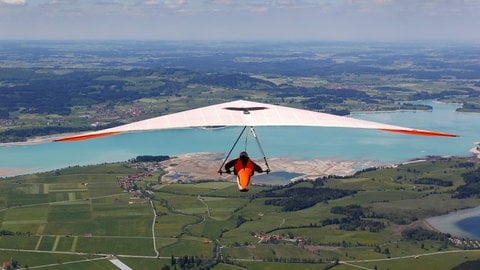 Ein weißer Drachen mit orangenem Innensegel - ein Drachenflieger schwebt in Richtung Forggensee über die Allgäuer Landschaft (Symbolbild). 