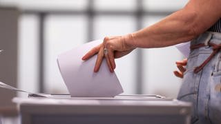 Eine Hand steckt Wahlunterlagen in eine Wahlurne. Am Sonntag werden bei der OB-Wahl 2023 die Ulmerinnern und Ulmer an die Wahlurnen gebeten, um eine neue Oberbürgermeisterin oder einen Oberbürgermeister zu wählen (Symbolbild). 