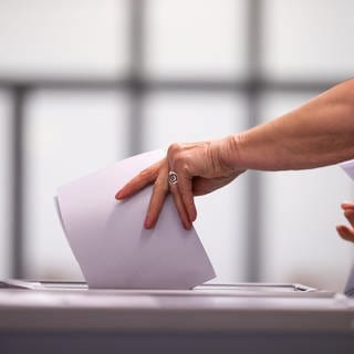 Eine Hand steckt Wahlunterlagen in eine Wahlurne. Am Sonntag werden bei der OB-Wahl 2023 die Ulmerinnern und Ulmer an die Wahlurnen gebeten, um eine neue Oberbürgermeisterin oder einen Oberbürgermeister zu wählen (Symbolbild). 