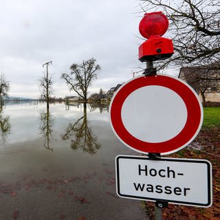 Hinter einem Durchfahrt verboten und Hochwasser-Hinweisschild, stehen Bäume im Wasser. Die Hochwasserlage bleibt in Teilen Baden-Württembergs weiterhin angespannt.
