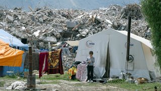 Junge steht vor einem Notunterkunft-Zelt, hinter ihm Berge von Schutt: Die Stadt Aalen (Ostalbkreis) will den Bau einer Rehaklinik in der türkischen Partnerstadt Antakya mitfinanzieren, die vor einem Jahr von einem Erdbeben betroffen war.