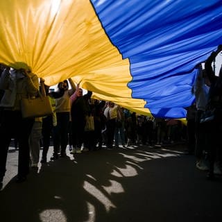 Demonstrierende halten eine große Ukraine-Flagge hoch, das Sonnenlicht scheint von oben hindurch. Der Angriffskrieg auf die Ukraine jährt sich bereits zum zweiten mal. Seitdem werden sowohl Friedensaktivisten als auch Rüstungshersteller in einem andern Licht gesehen.