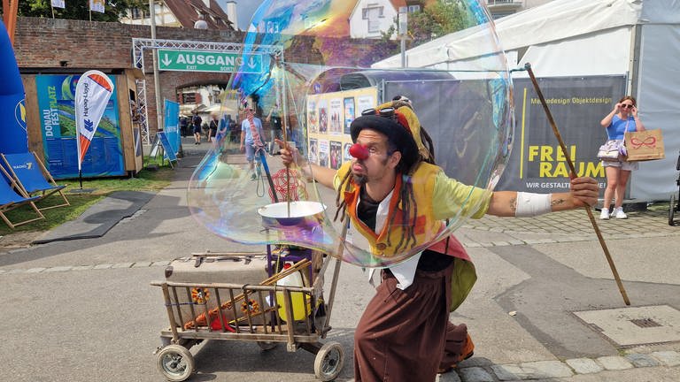 Ein Clown bewegt eine Riesenseifenblase mit zwei langen Holzstäben in der Luft - ein Hingucker auf dem Donaufest am Ulmer Ufer.  