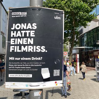Ein schwarzes Plakat mit weißer Schrift das auf das Pilotprojekt, Ulm testet auf K.O. hinweist. Eines von mehreren Plakaten, die in Ulm auf K.o.-Tropfen aufmerksam machen soll. Wer es mit dem Handy fotografiert, wird auf eine Aufklärungsseite der Stadt Ulm weitergeleitet.
