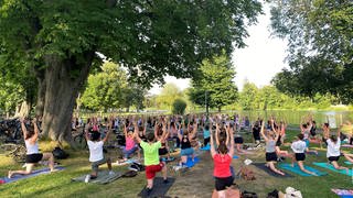 Ein Yogakurs in einem Park. Sport machen: Gemeinsam draußen und das auch noch kostenlos, das geht im Sommer in Ulm. Von Mai bis September bietet die Stadt Ulm kostenlose Sportkurse  wie Tanzen, Fitness, Selbstverteidigung in mehreren Parks an, und das sehr erfolgreich. Zu "Yoga auf Englisch" kommen oft mehr als 100 Teilnehmende.