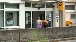 Zwei Frauen stehen mit einem Paket vor der Post in Blaustein. Sie lesen ein Schild, das auf die Schließung hinweist: Die Postagentur im Zentrum Blausteins ist geschlossen.