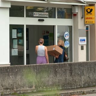 Zwei Frauen stehen mit einem Paket vor der Post in Blaustein. Sie lesen ein Schild, das auf die Schließung hinweist: Die Postagentur im Zentrum Blausteins ist geschlossen.