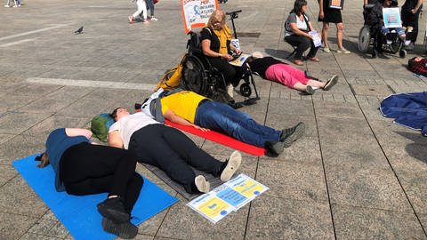 Mehrere Menschen liegen auf dem Münsterplatz in Ulm auf dem Boden. Mit der Liegend-Demo wollen sie auf das chronische Fatigue Syndrom oder auch MECFS aufmerksam machen und fordern eine bessere Behandlung.