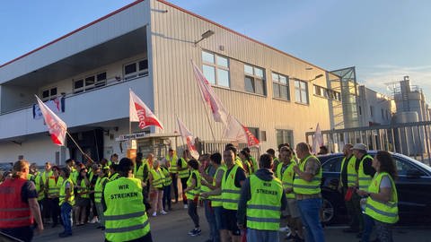 Streikende mit signalgelben Westen und Fahnen stehen vor der Lebkuchenfabrik Max Weiss in Neu-Ulm.