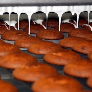 Runde Lebkuchen fahren auf einem Produktionsband unter eine Maschine, die weiße Glasur drauftropft (Archivbild). Allerdings produziert die Lebkuchenfirma Weiss in Neu-Ulm derzeit nicht: Alle drei Produktionsschichten sind am Mittwoch zum Warnstreik aufgerufen.