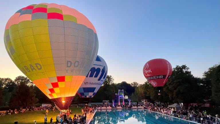 Viele Menschen verfolgen am Schwimmerbecken im Donaubad Neu-Ulm die Taufe eines erleuchteten Heißluftballons.