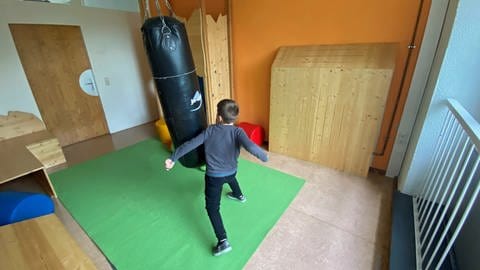 Ein Junge boxt gegen einen Boxsack. Er hängt in einem bunt eingerichteten Zimmer mit Kletterwand und Schaumstoffmatten. Hier können sich die Kinder bei Bedarf austoben. 