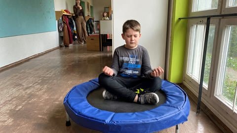 Ein Junge sitzt mit geschlossenen Augen im Schneidersitz auf einem kleinen Trampolin in einem Schulhaus-Gang und meditiert. Die Hans-Zulliger-Schule in Ulm ist für Kinder, die keine "normale" Grundschule besuchen können.