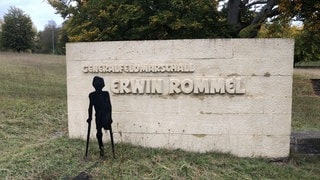 Eine mit einer Krücke gehende Figur steht am 80. Todestag Rommels vor einem großen, weißen Gedenkstein in Heidenheim mit der Inschrift "Generalfeldmarschall Erwin Rommel".