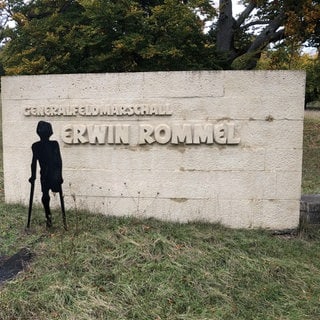 Eine mit einer Krücke gehende Figur steht am 80. Todestag Rommels vor einem großen, weißen Gedenkstein in Heidenheim mit der Inschrift "Generalfeldmarschall Erwin Rommel".